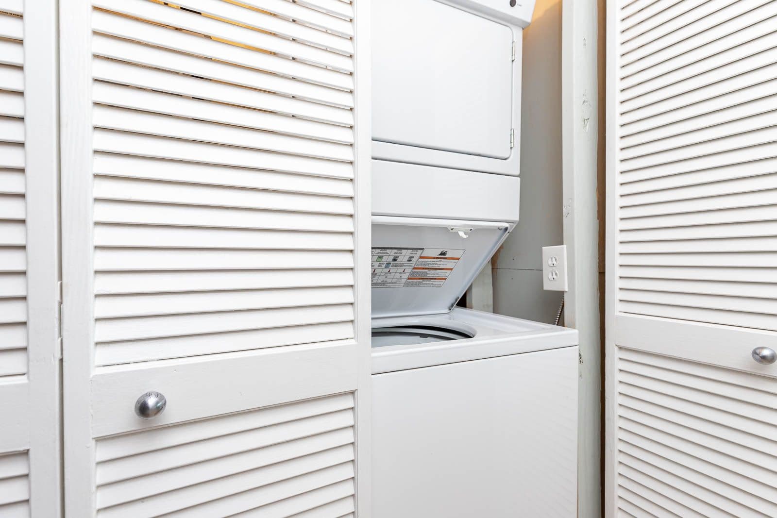 Une petite buanderie blanche, dotée d’un lave-linge et d’un sèche-linge intégrés, et d’un panneau de commande avec instructions.