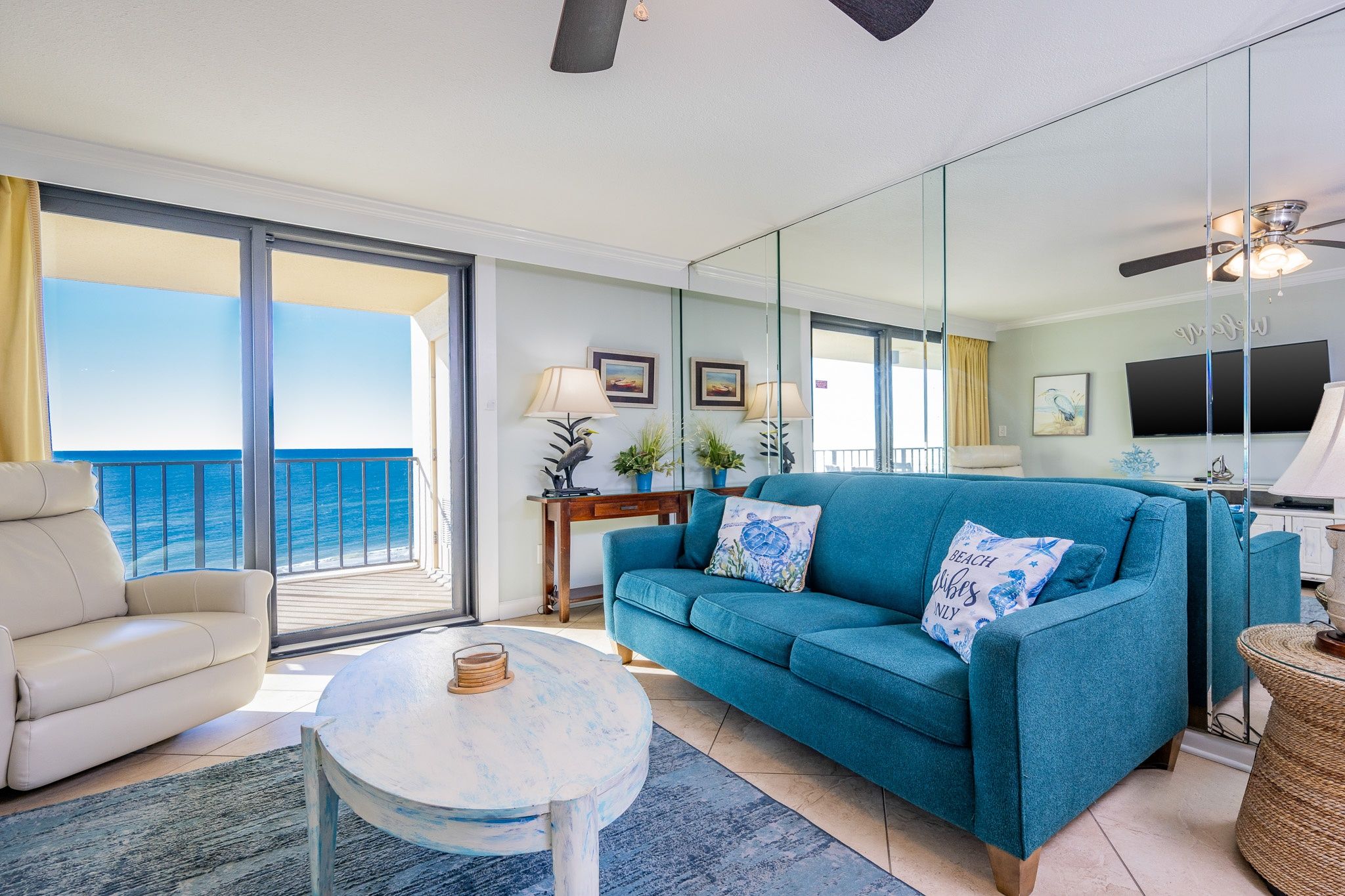 A living room with a sofa, armchair, and round coffee table, a balcony with a view of the sea, and a television.