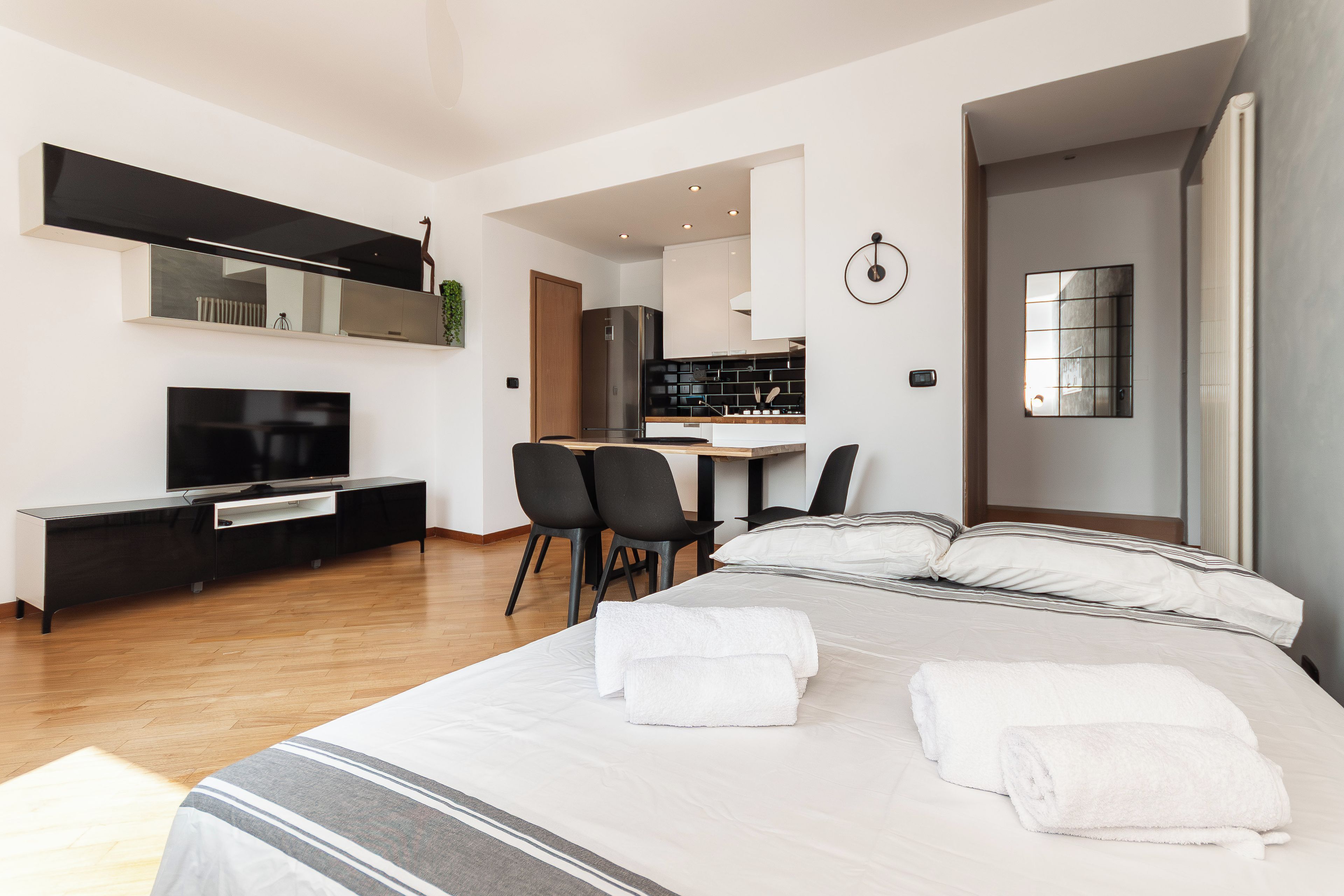A modern hotel room with a large bed, a TV, a desk, and a kitchen area.
