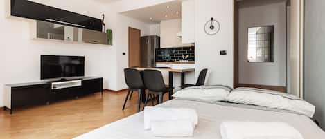 A modern hotel room with a large bed, a TV, a desk, and a kitchen area.