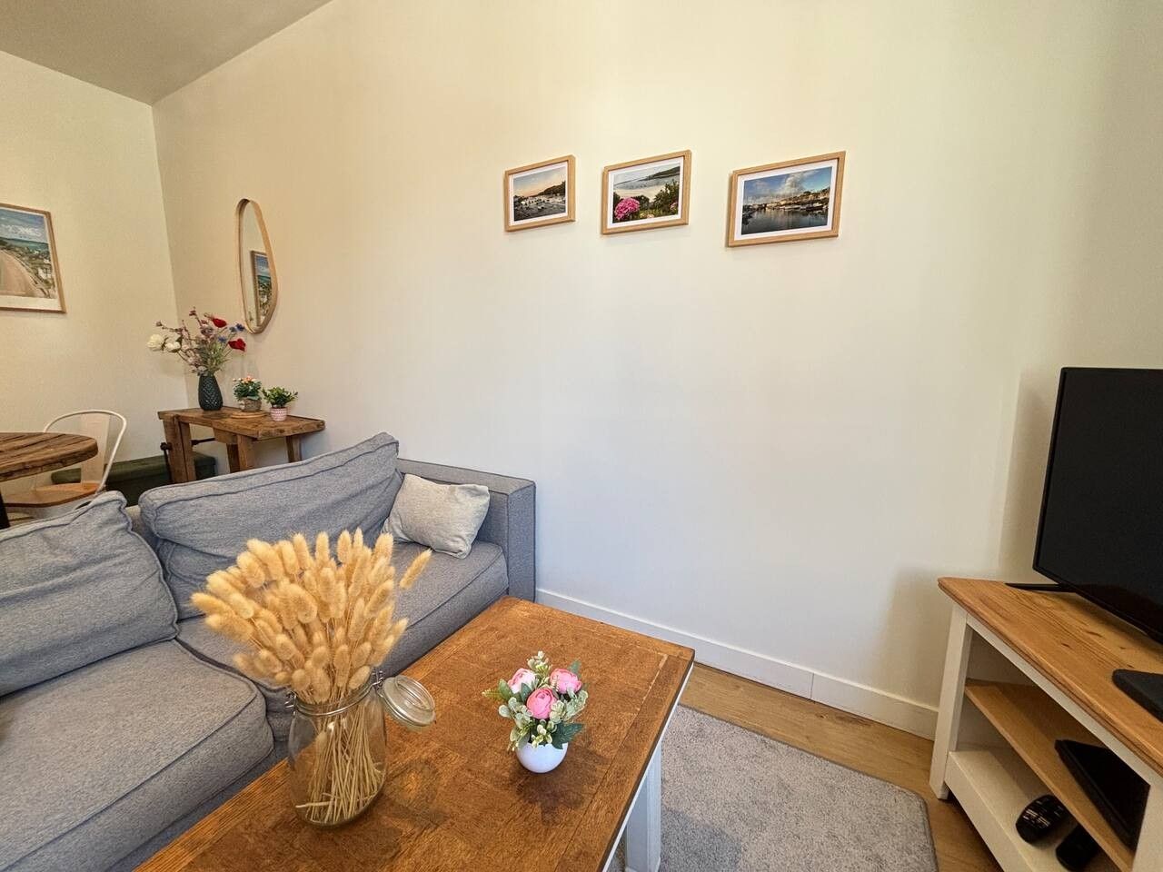 A living room with a grey sofa, a wooden coffee table with flowers, a TV stand, and three framed pictures on the wall.