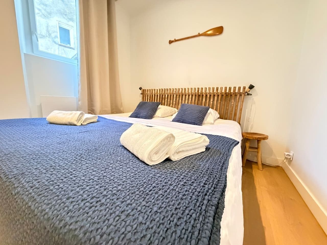 Una cama bien hecha con una manta azul y dos toallas enrolladas, un cabecero de madera, un taburete pequeño de madera y una pala montada en la pared.