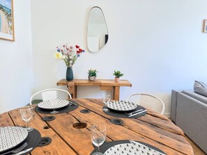 Ein Holztisch gedeckt mit Tellern, Besteck und Weingläsern, ein Spiegel an der Wand und eine kleine Vase mit Blumen auf einem Beistelltisch.