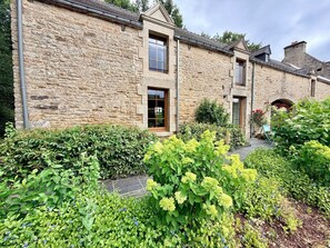 A stone building with a wooden door and windows, surrounded by a well-maintained garden with various plants and shrubs. - Charming Vacation Rental - Stone and Wood - Morbihan (Saint-Jean-Brévelay)
