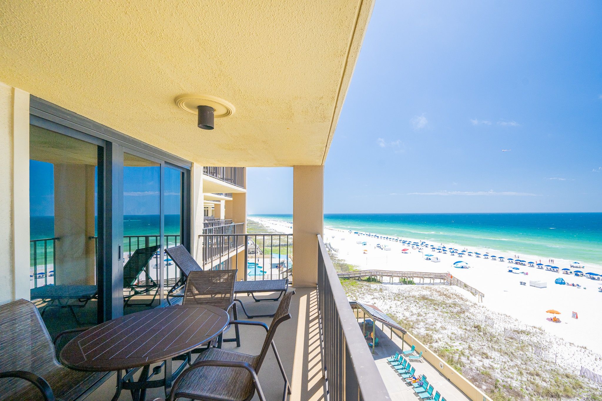 A balcony with a table and chairs, overlooking a beach with umbrellas and lounge chairs.