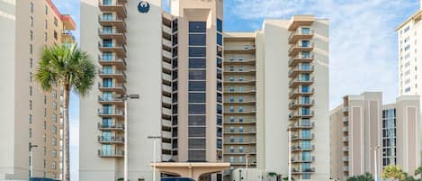 Um hotel com vários andares, uma torre central, varandas e um estacionamento com diversos veículos.