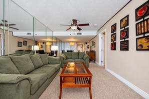 A living room with a green sofa, a wooden coffee table, a ceiling fan, and wall-mounted artwork. - Phoenix I 1012 2 Bedroom Condo (Orange Beach)