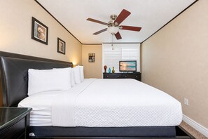 Una habitación de hotel con cama, mesita de noche, televisión, ventilador de techo y obras de arte colgadas en la pared.