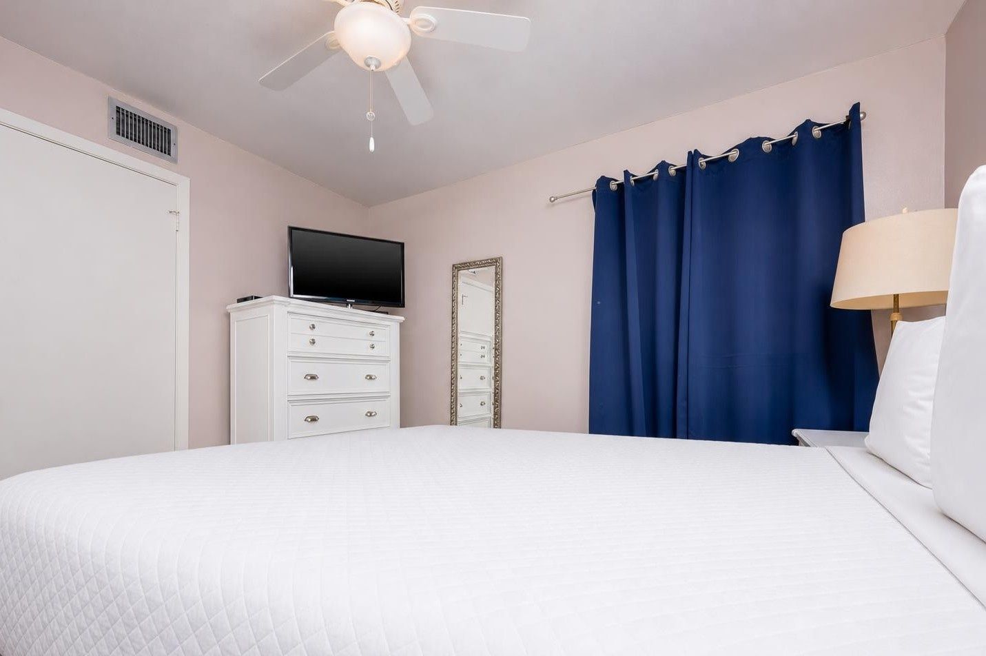 Une chambre à coucher comprenant un lit, une commode, un téléviseur, un ventilateur de plafond et une fenêtre avec des rideaux.