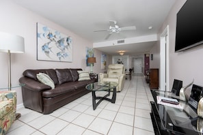 A living room with a brown leather sofa, a glass coffee table, a beige armchair, and a flat-screen TV mounted on the wall. - Phoenix I 1073 2 Bedroom Condo (Orange Beach)