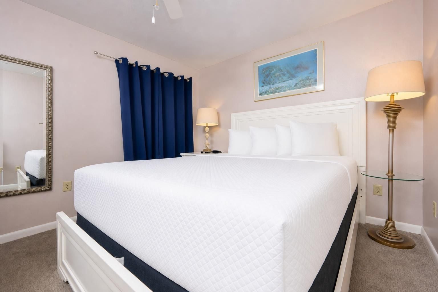 A hotel room with a large bed, a nightstand, a lamp, a mirror, and a painting on the wall.