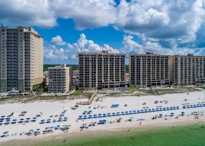 A beachfront hotel with multiple high-rise buildings, a sandy beach with numerous umbrellas, and a clear blue sky. - Phoenix I 1073 2 Bedroom Condo (Orange Beach)