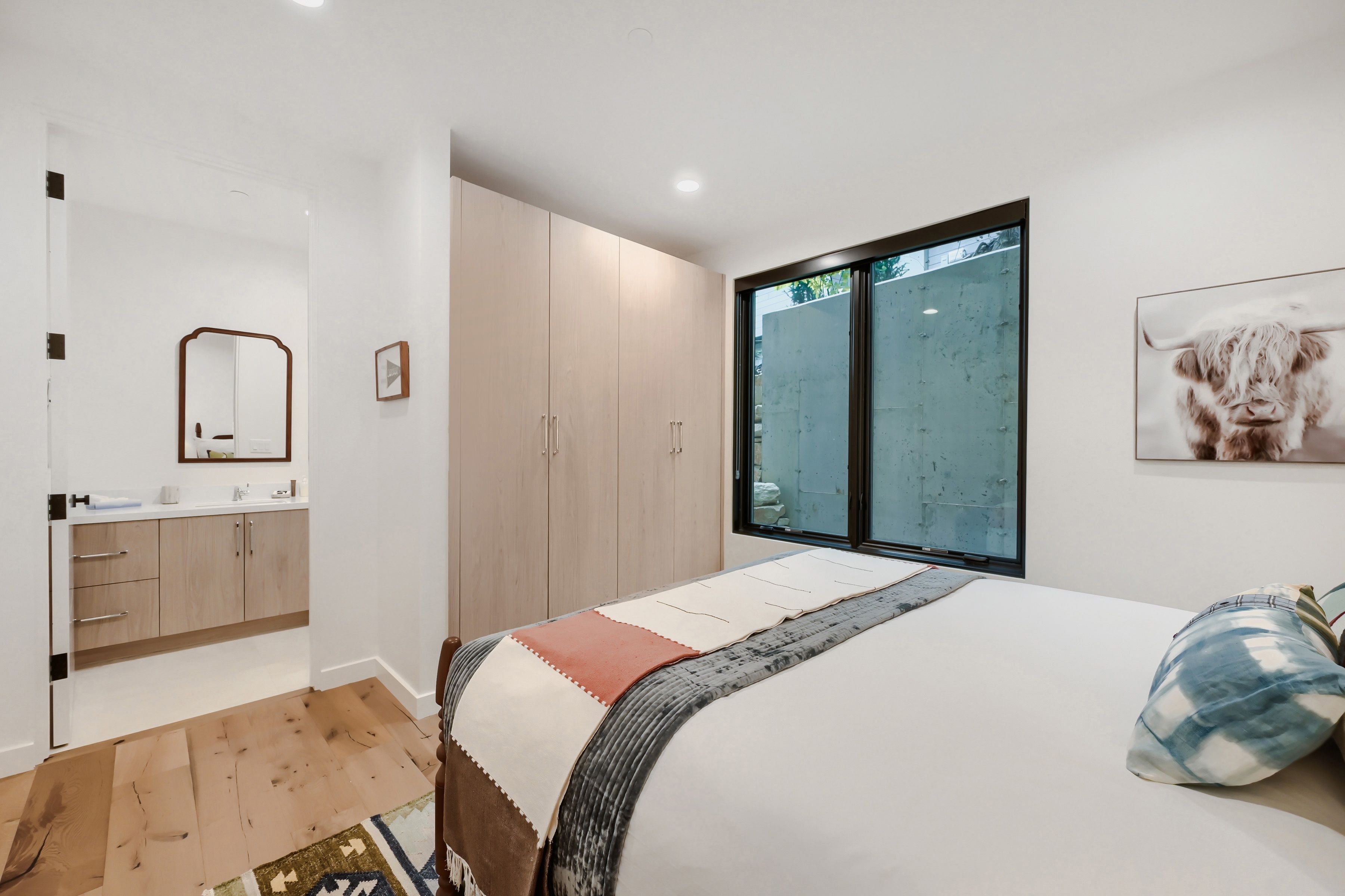 A bedroom with a large window, a bed with a patterned bedspread, a bedside table, and a wardrobe.