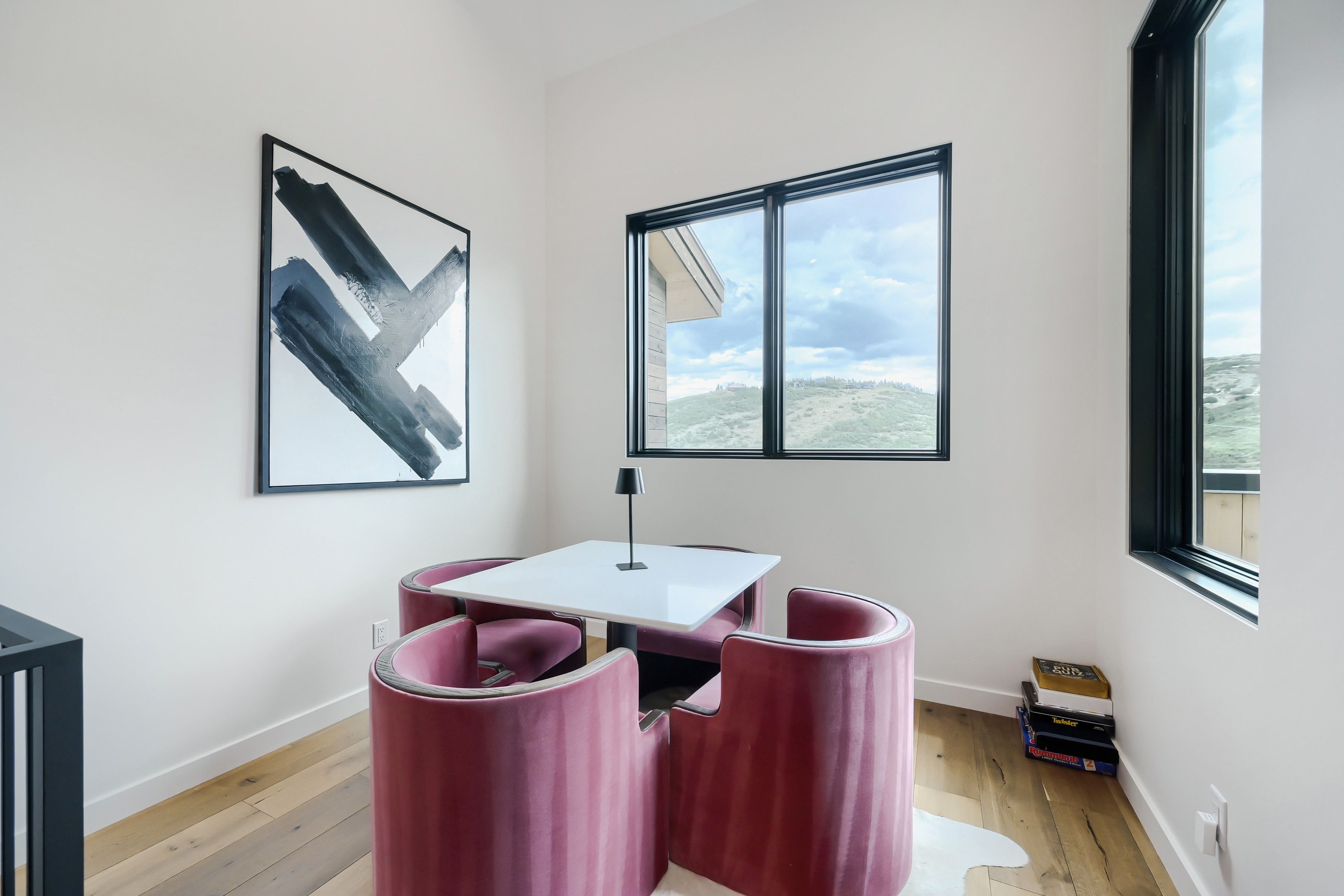 Een moderne kamer met een witte tafel, twee roze stoelen, een abstract schilderij in het zwart, een raam met uitzicht op een glooiend landschap, en een lamp.