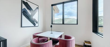 Een moderne kamer met een witte tafel, twee roze stoelen, een abstract schilderij in het zwart, een raam met uitzicht op een glooiend landschap, en een lamp.