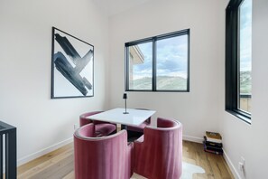 Ein modernes Zimmer mit einem weißen Tisch, zwei rosafarbenen Stühlen, einem schwarzen abstrakten Gemälde, einem Fenster mit Blick auf eine hügelige Landschaft und einer Lampe.