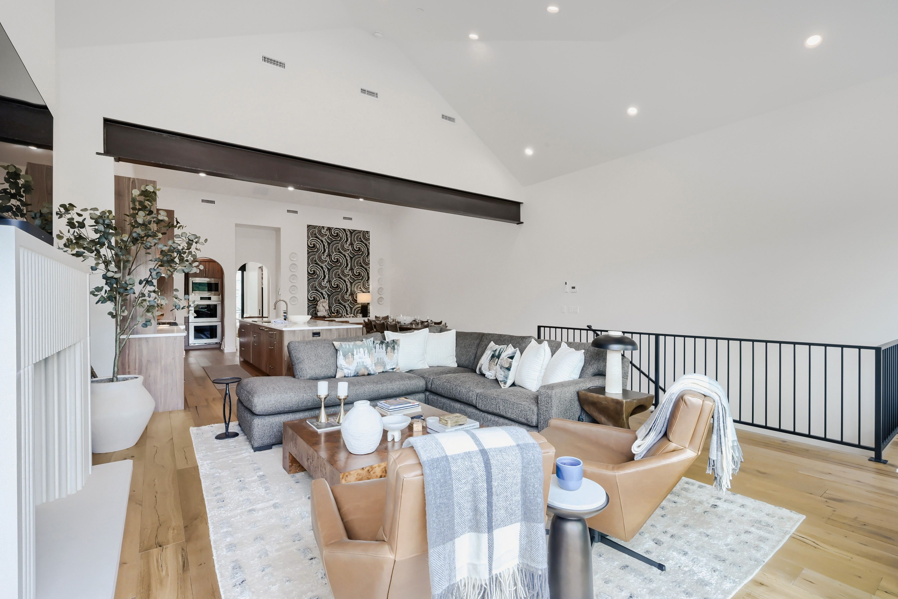 A modern living room with a grey sectional sofa, a coffee table with decorative items, and a view of the kitchen and dining area in the background.