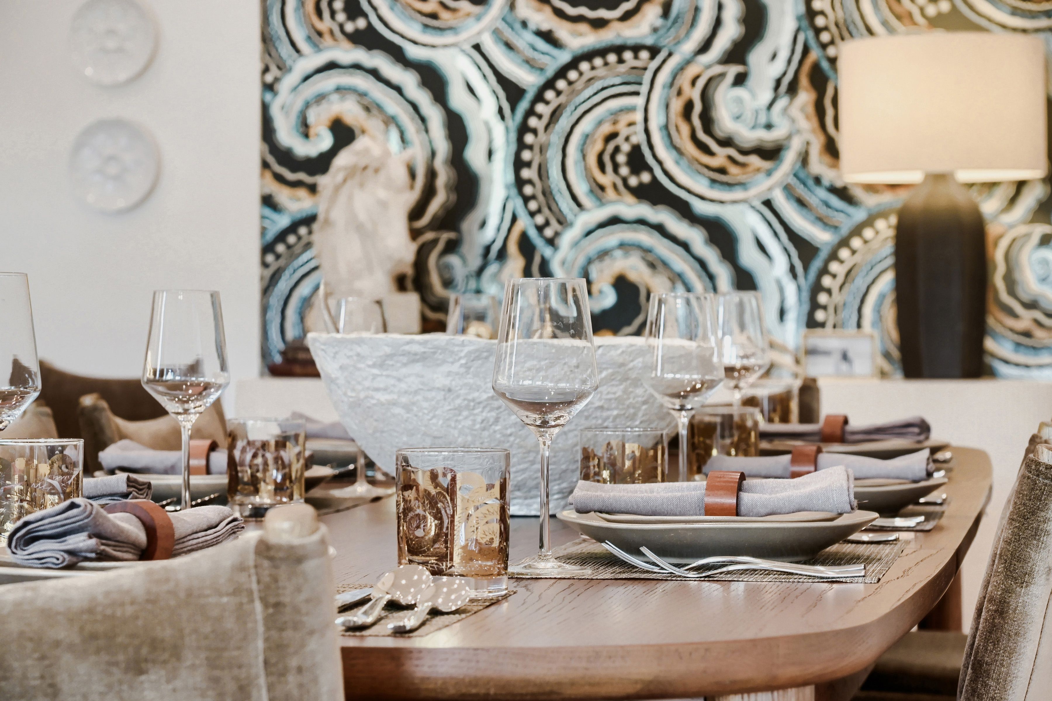 Mesa de comedor dispuesta con vasos, platos y cubiertos, con un papel tapiz estampado en el fondo.