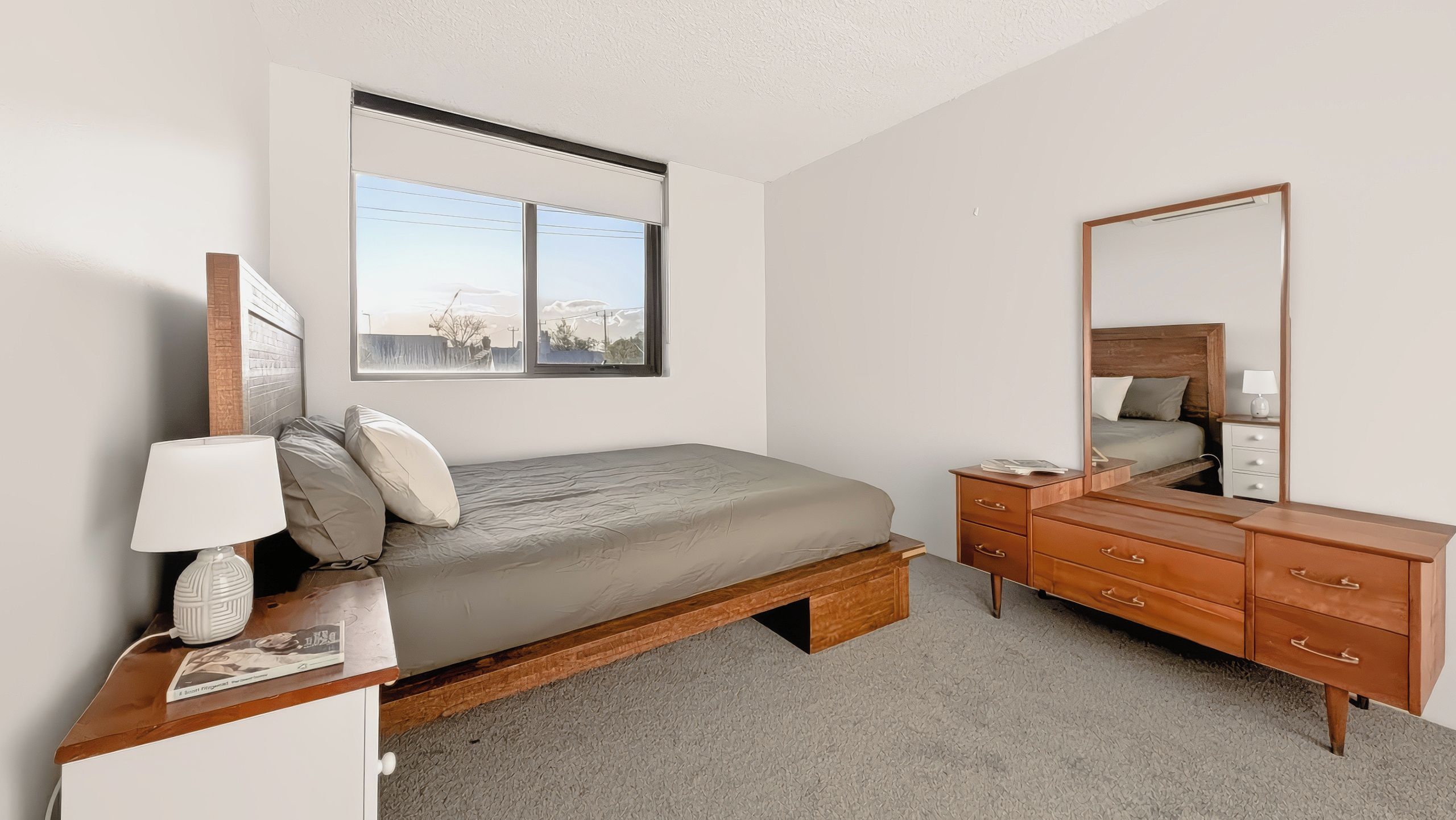 A bedroom with a bed, bedside table, lamp, dresser, and a large window.