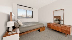 Quarto com cama, mesa de cabeceira, luminária, cômoda e uma janela grande.