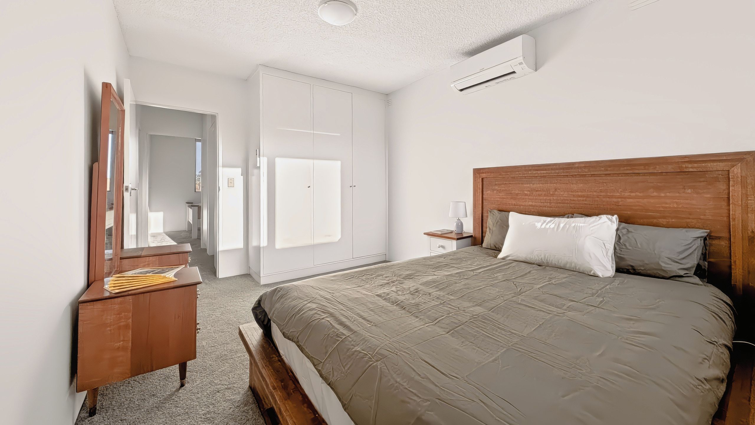 A bedroom with a bed, a nightstand with a lamp, a dresser, a mirror, and a door leading to another room.