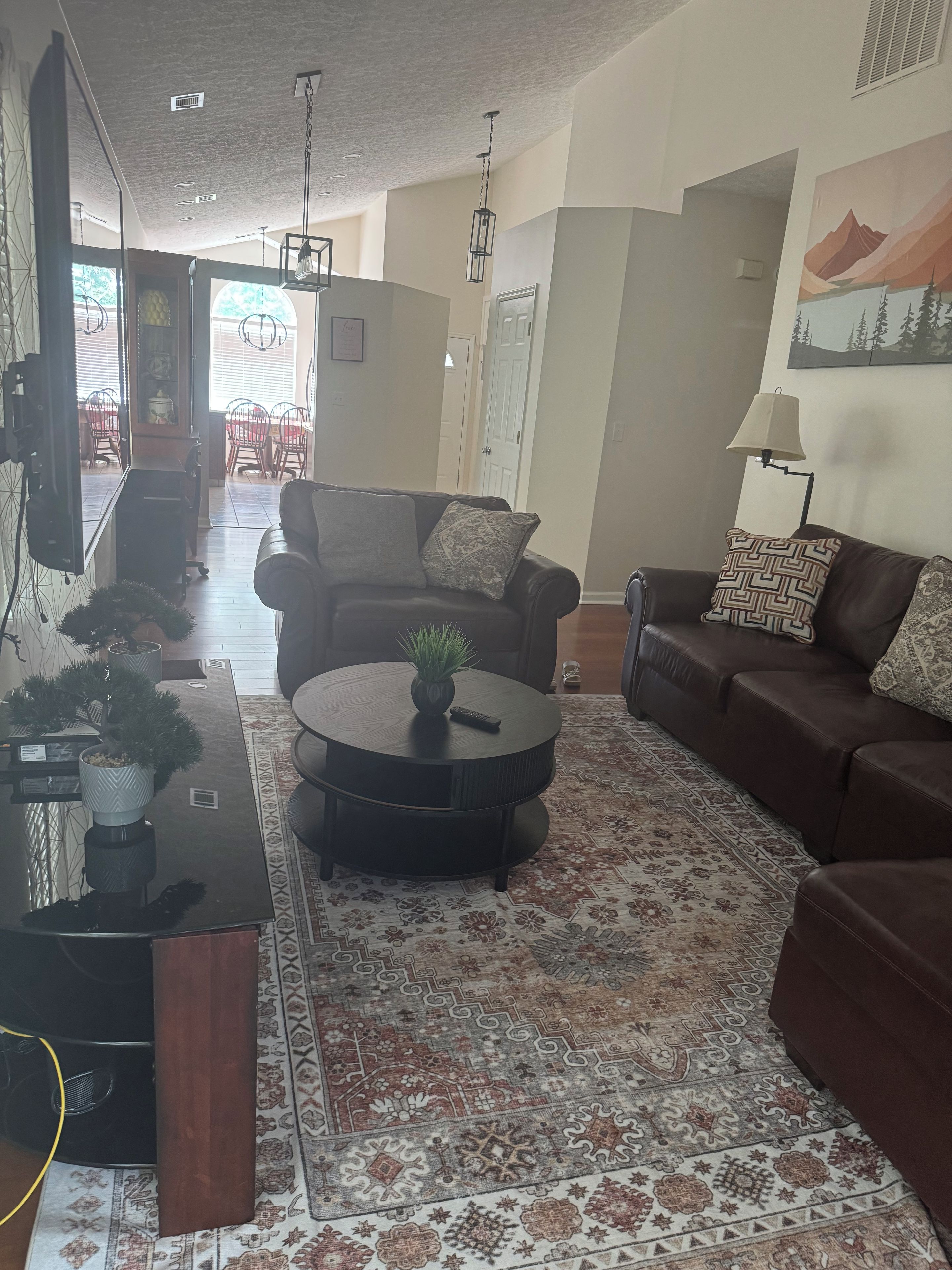 Uma sala de estar com um sofá de couro marrom, uma mesa de centro preta e um tapete estampado. Ao fundo, há uma área de jantar com uma mesa e cadeiras.
