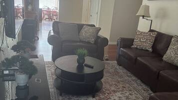 Uma sala de estar com um sofá de couro marrom, uma mesa de centro preta e um tapete estampado. Ao fundo, há uma área de jantar com uma mesa e cadeiras.