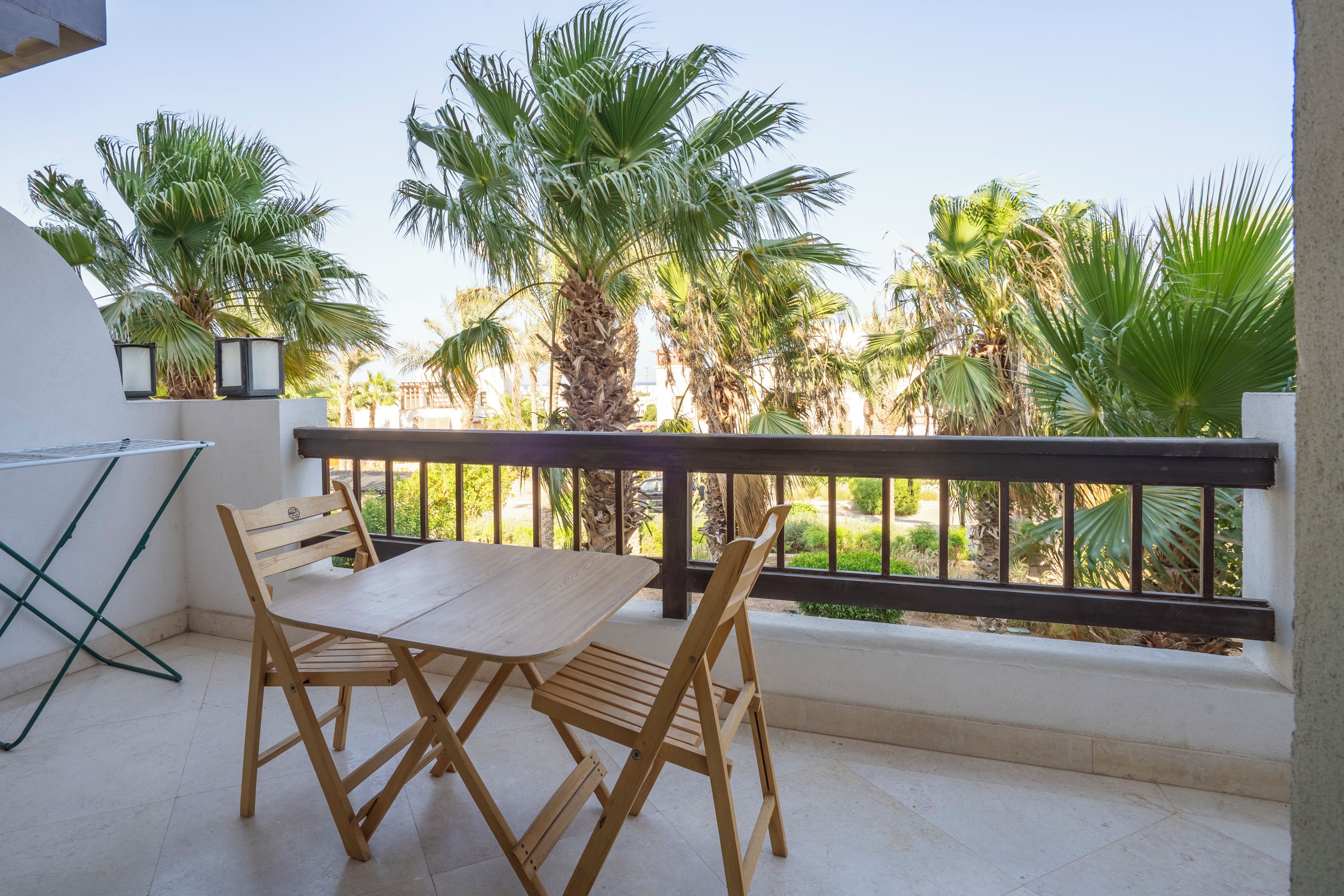 Un balcon avec une table et des chaises en bois, un barbecue en métal et une vue sur des palmiers et la végétation.