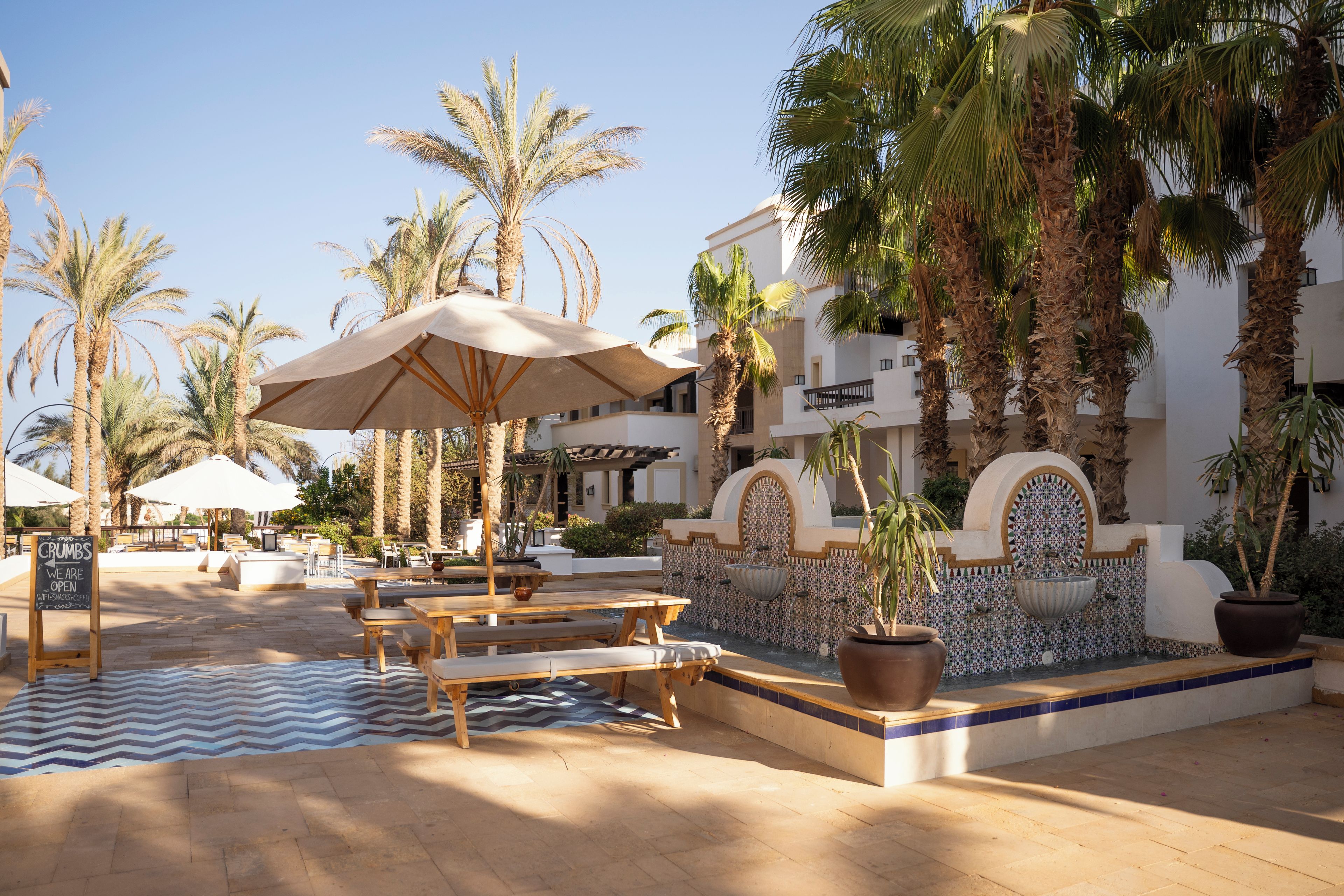 An outdoor seating area with tables and umbrellas, surrounded by palm trees and a building with tiled walls.