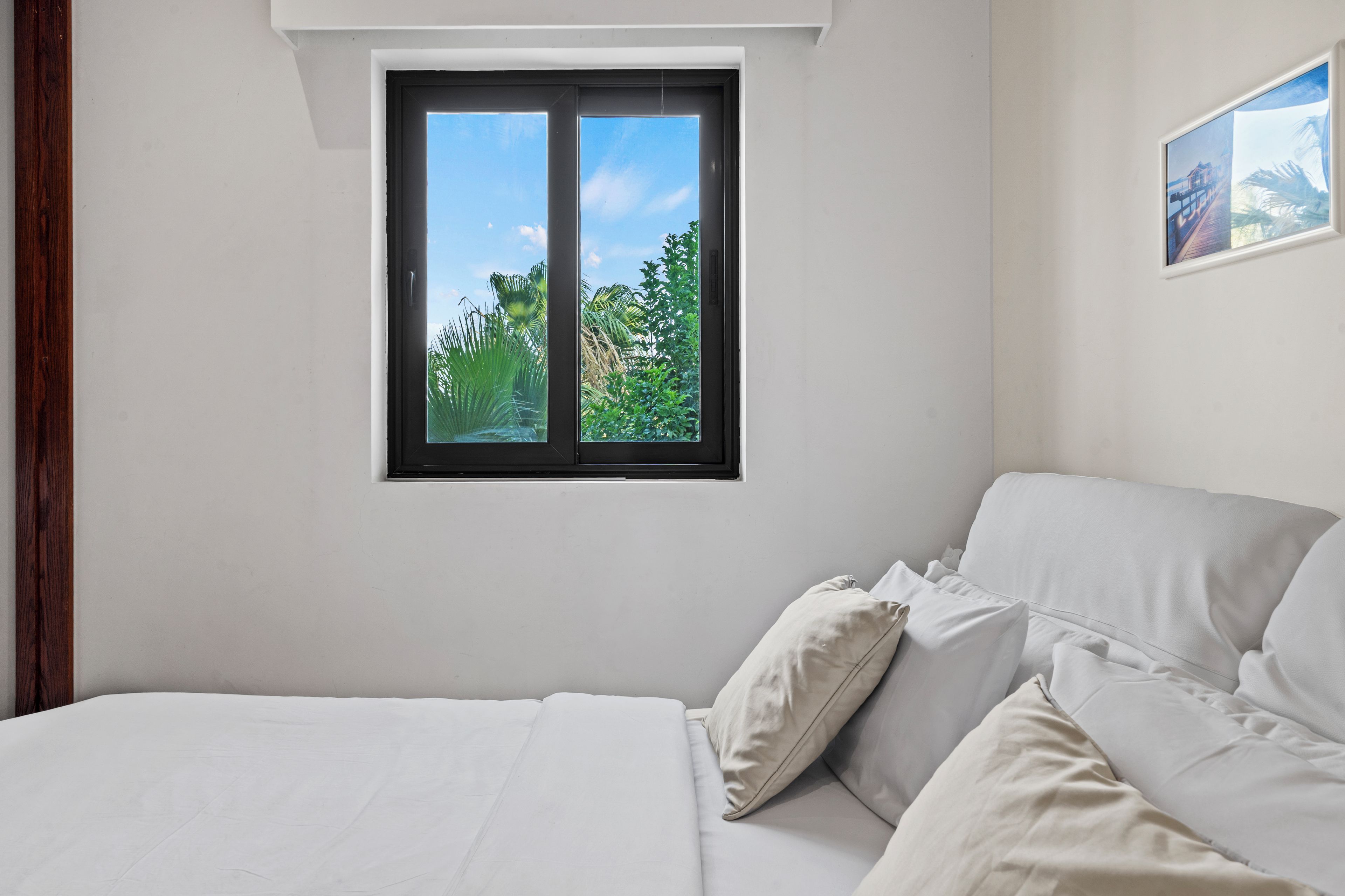 Une chambre à coucher avec un lit, des oreillers, une fenêtre donnant sur un paysage verdoyant et un tableau encadré accroché au mur.
