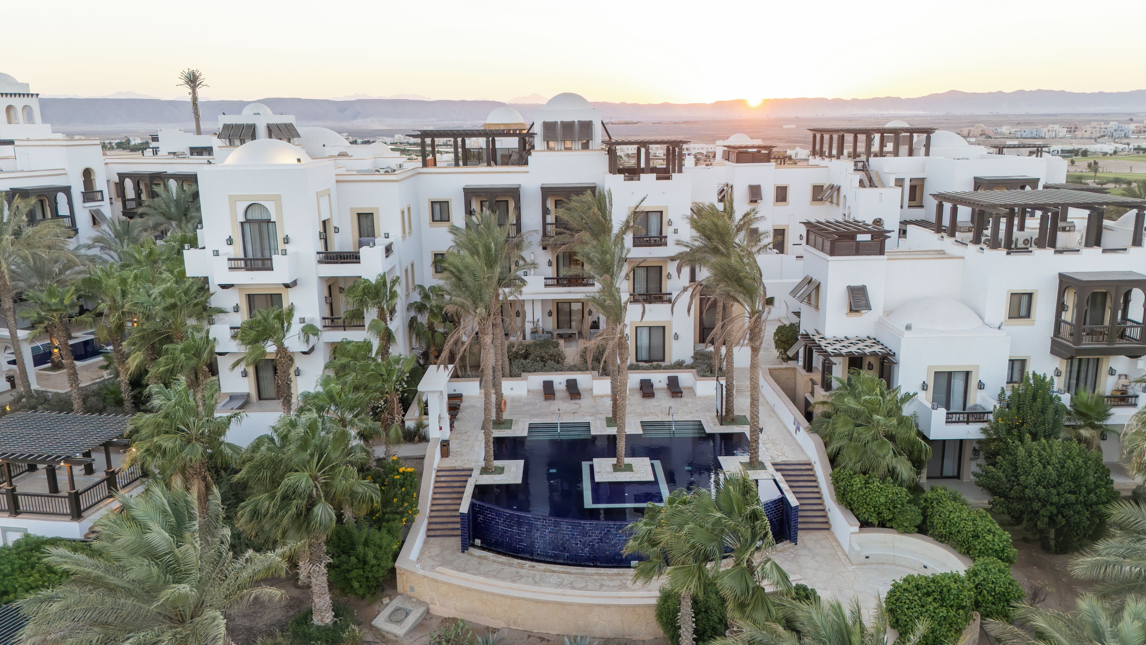 A resort with multiple white buildings, palm trees, and a central swimming pool.