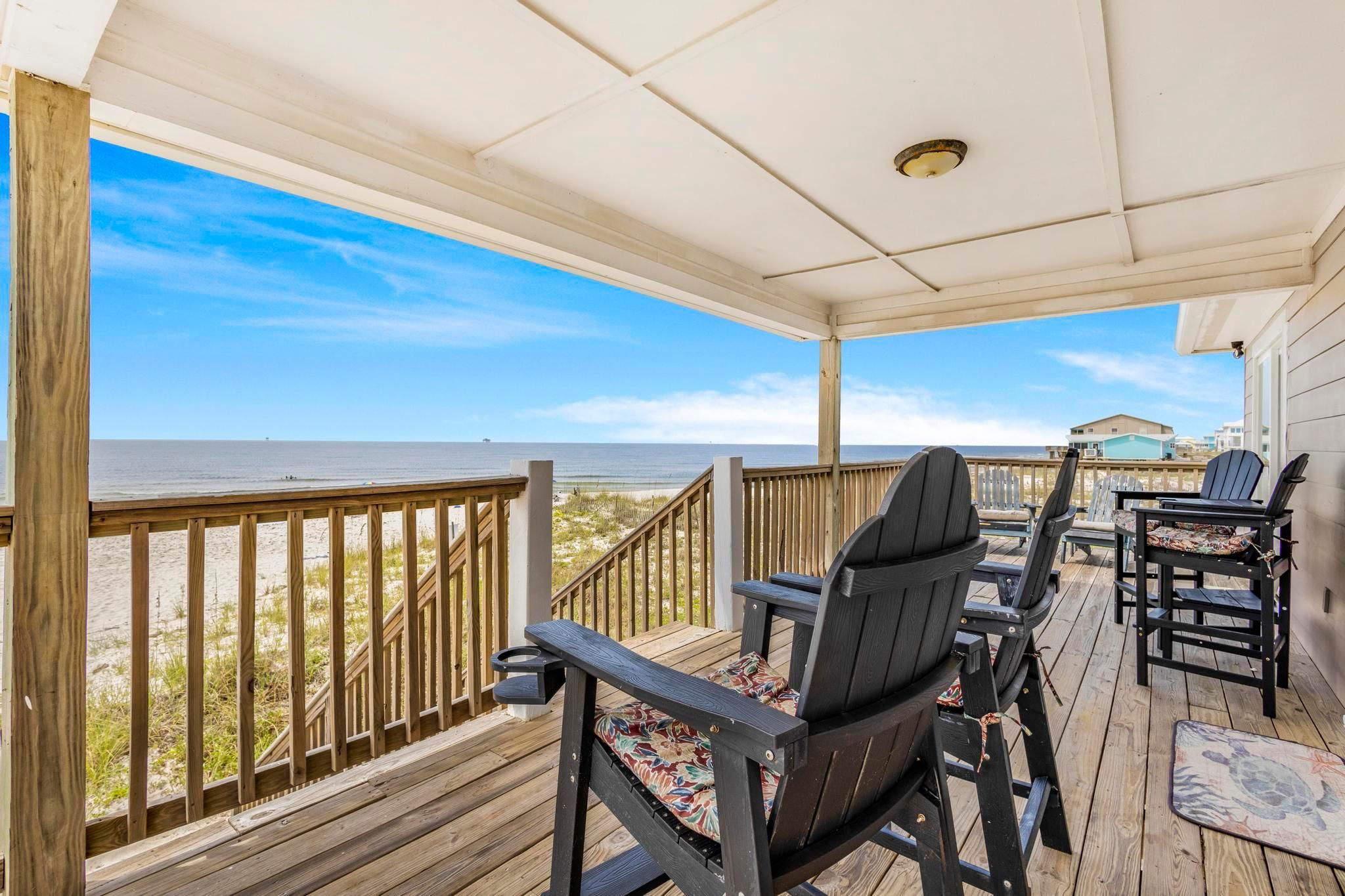 A covered outdoor deck with wooden railings, a view of the beach, and several Adirondack chairs.