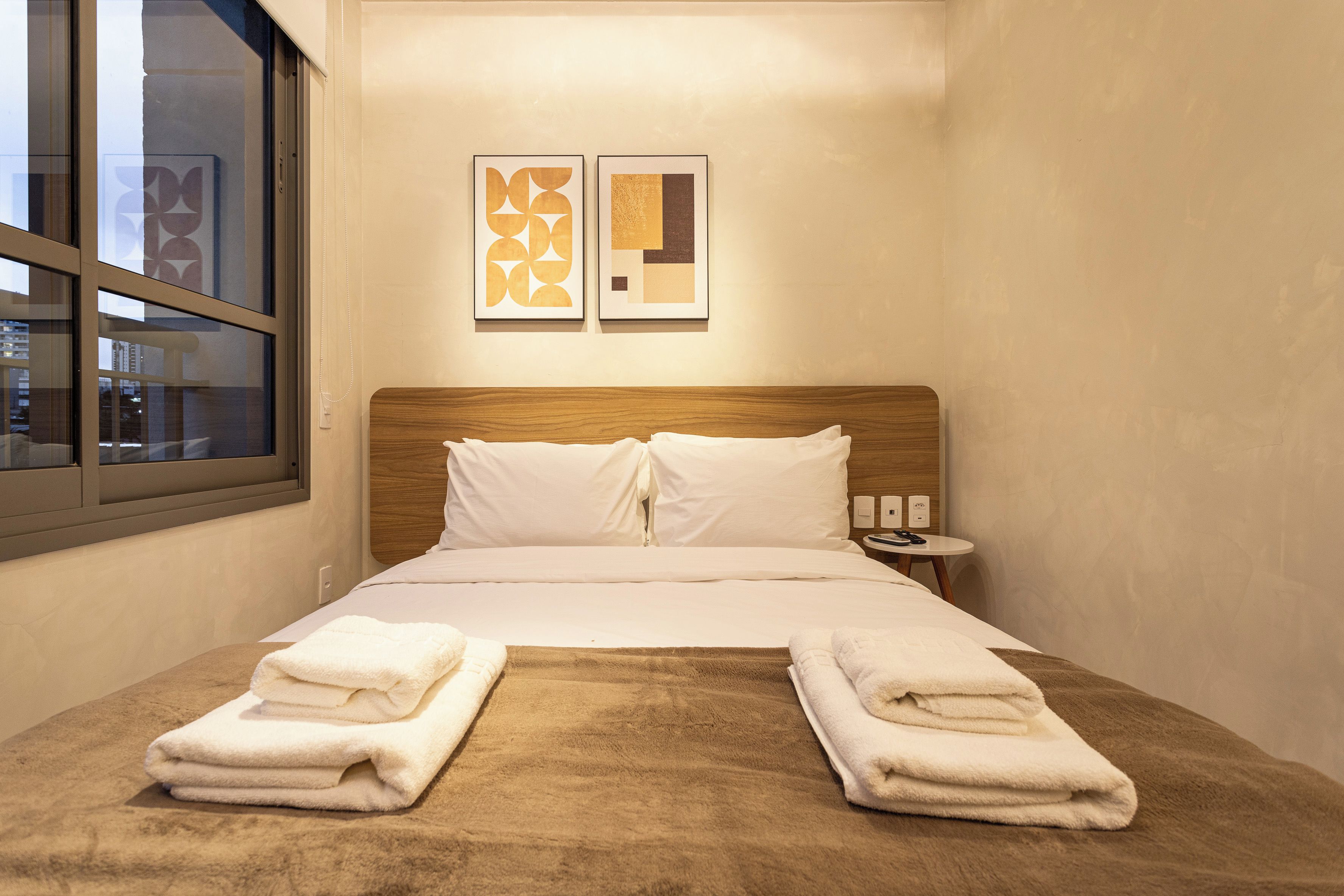 A hotel room with a bed, two pillows, a wooden headboard, a nightstand with a lamp, and two framed artworks on the wall.