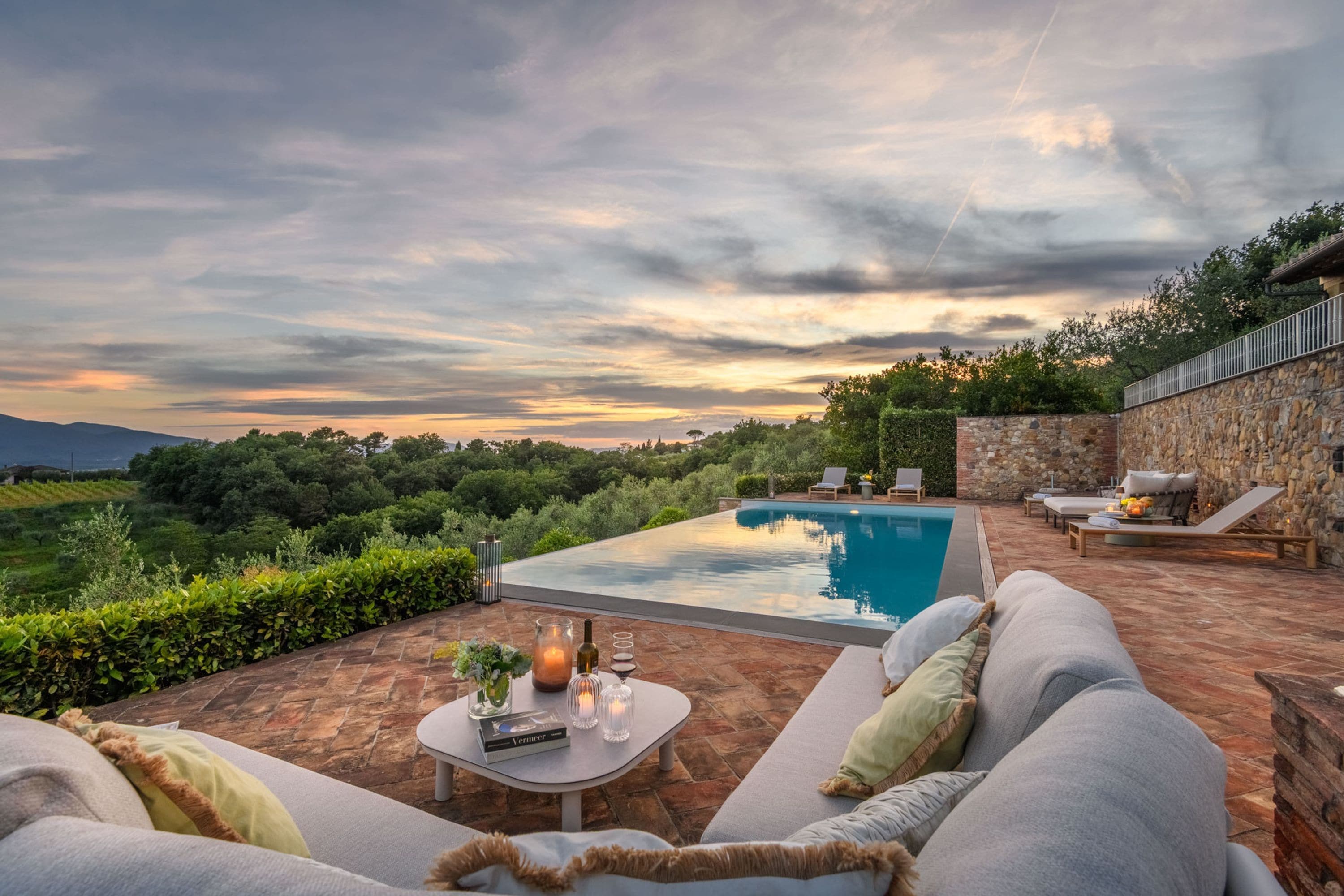A poolside area with a stone wall, outdoor seating, and a view of the landscape.