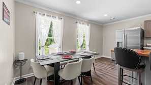 A dining area with a table set for four, chairs, a small side table, and a kitchen area with a refrigerator. - 4BR Gameday Home + Separate Guest Suite (Tuscaloosa)