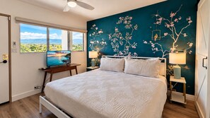 A bedroom with a large bed, a TV on a stand, a window with a view, and a wall with a floral mural. - Where Paradise Meets the Waves Remodeled Kihei Condo with Ocean Views (Kihei)