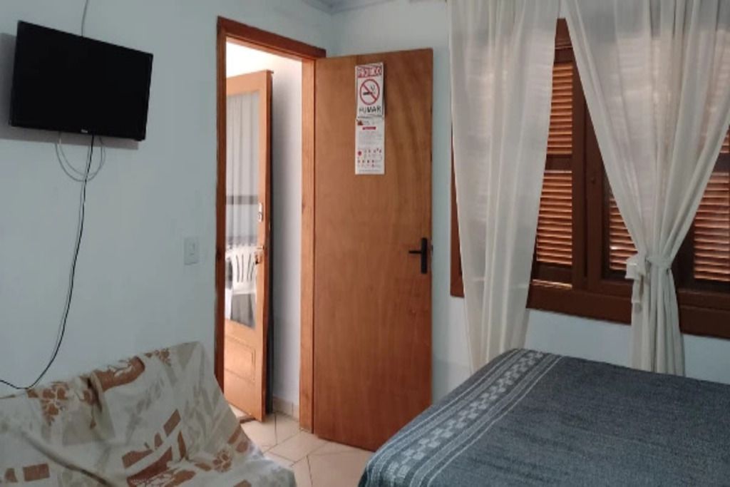 Quarto com cama, televisão fixada na parede, porta com placa e janela com cortinas.