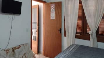 Quarto com cama, televisão fixada na parede, porta com placa e janela com cortinas.