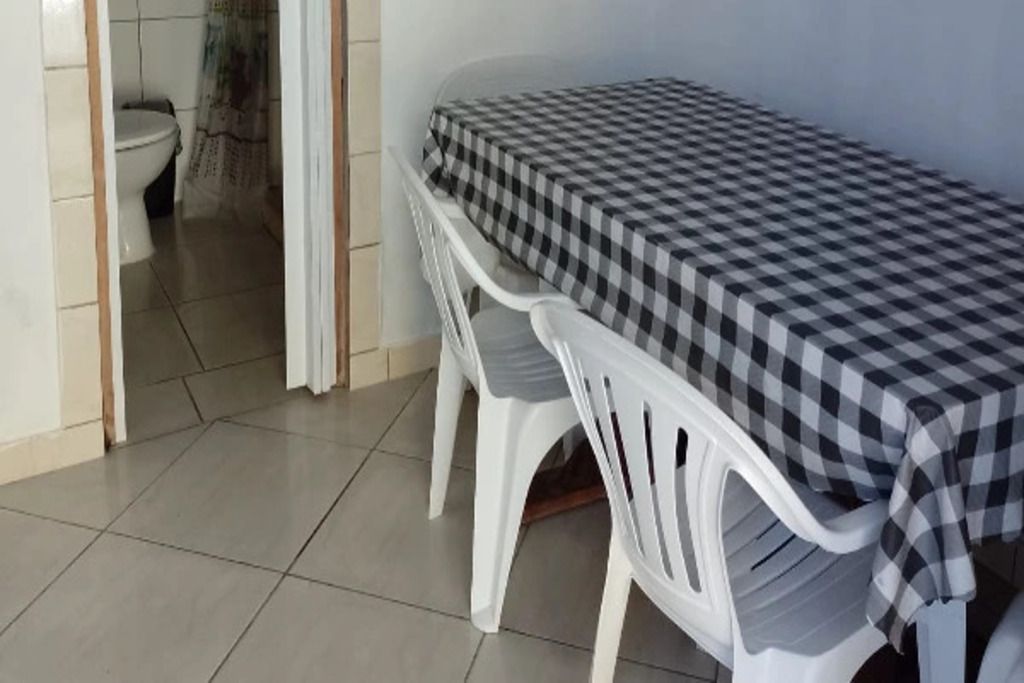 Quarto pequeno com uma mesa xadrez e duas cadeiras de plástico brancas. Um sanitário é visível ao fundo.
