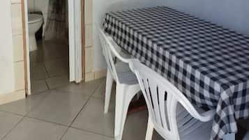 Quarto pequeno com uma mesa xadrez e duas cadeiras de plástico brancas. Um sanitário é visível ao fundo.