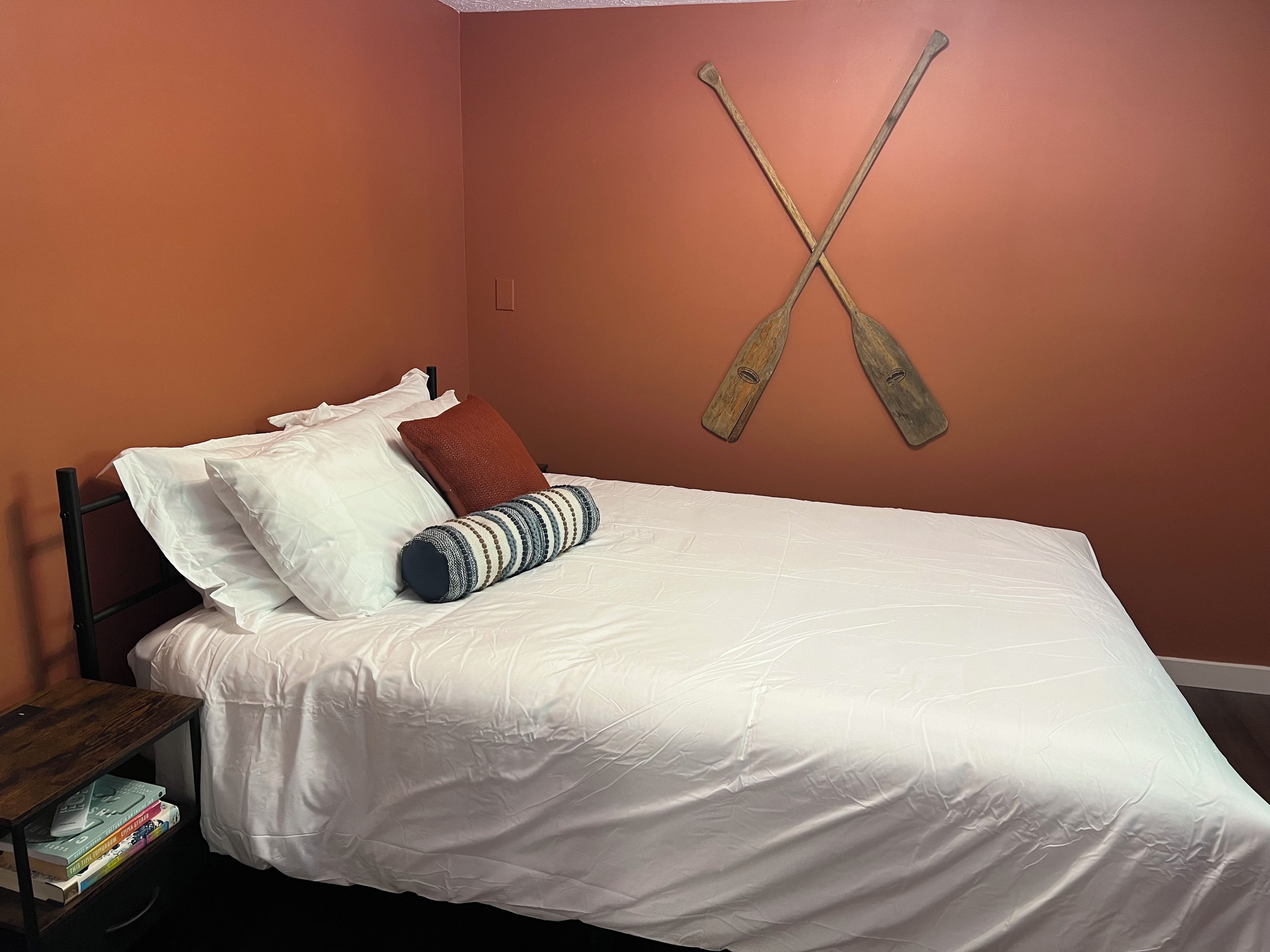 Cama com roupa de cama branca, travesseiro listrado e travesseiro vermelho. Tem uma mesa de cabeceira de madeira com livros e uma luminária. A parede é laranja, e dois remo de madeira estão pendurados nela.