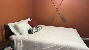 Cama com roupa de cama branca, travesseiro listrado e travesseiro vermelho. Tem uma mesa de cabeceira de madeira com livros e uma luminária. A parede é laranja, e dois remo de madeira estão pendurados nela.