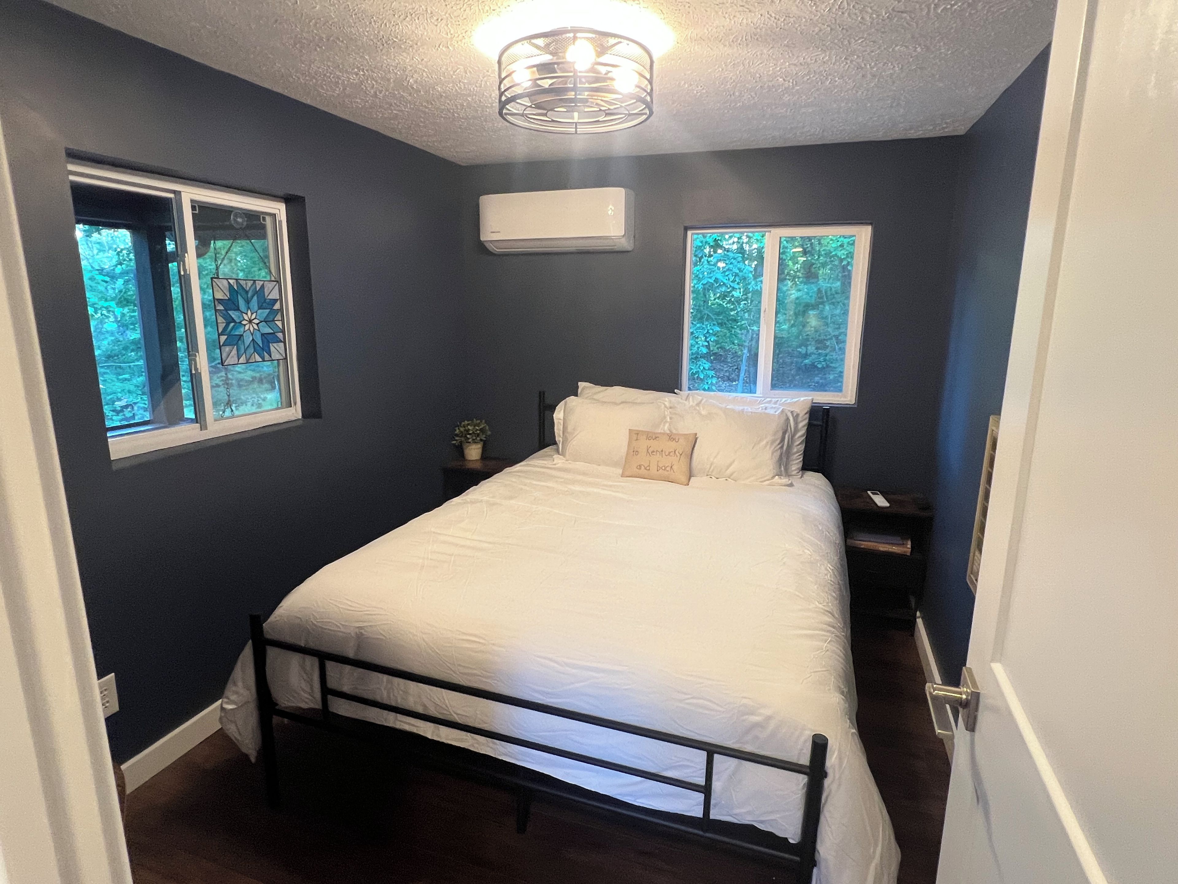 Un dormitorio con una cama, dos ventanas, una lámpara de techo y un aire acondicionado de pared.