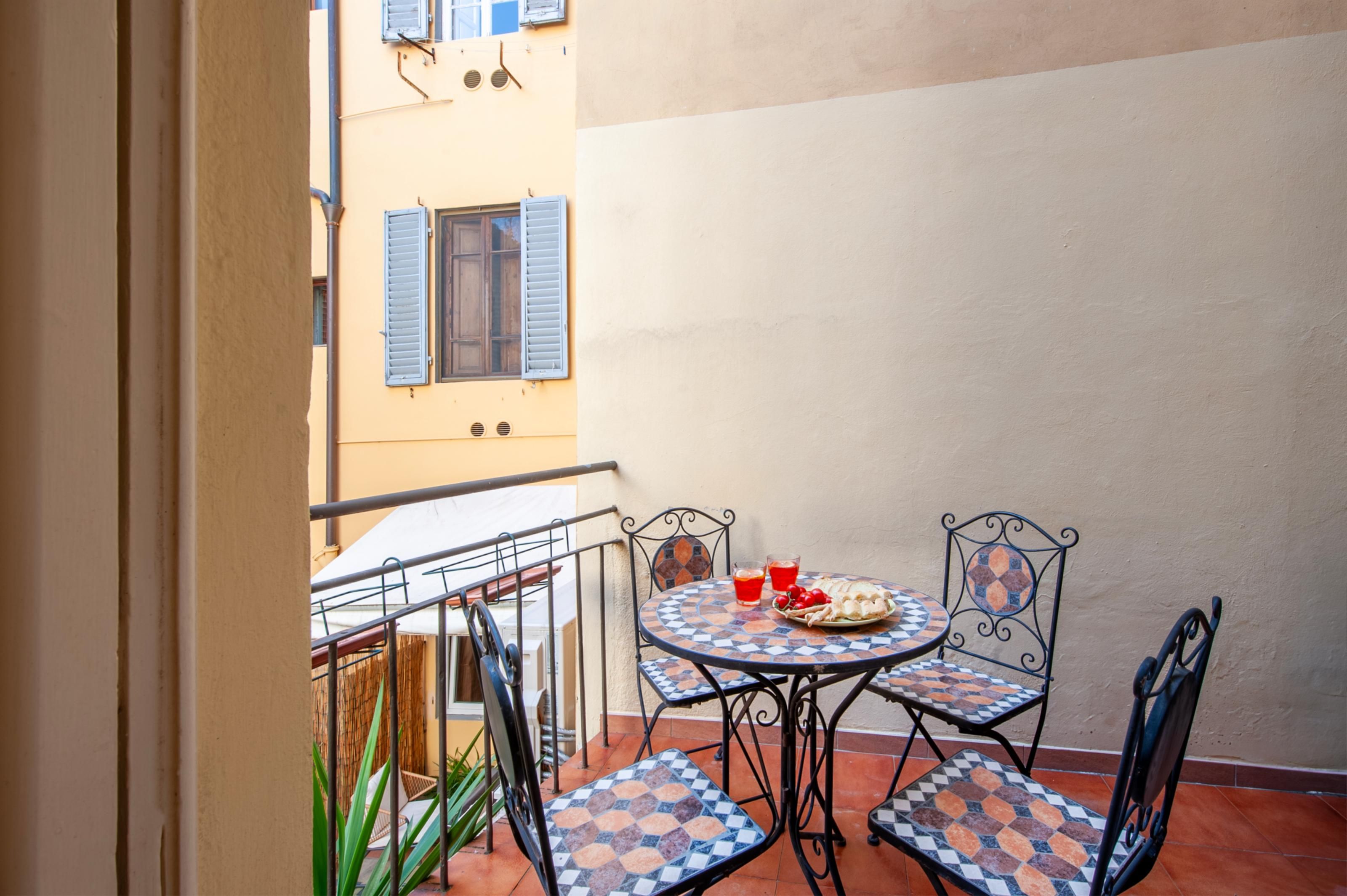 Un pequeño patio al aire libre con una mesa redonda y cuatro sillas, una mesita con un plato de comida y bebidas, y vistas a un edificio con contraventanas azules.
