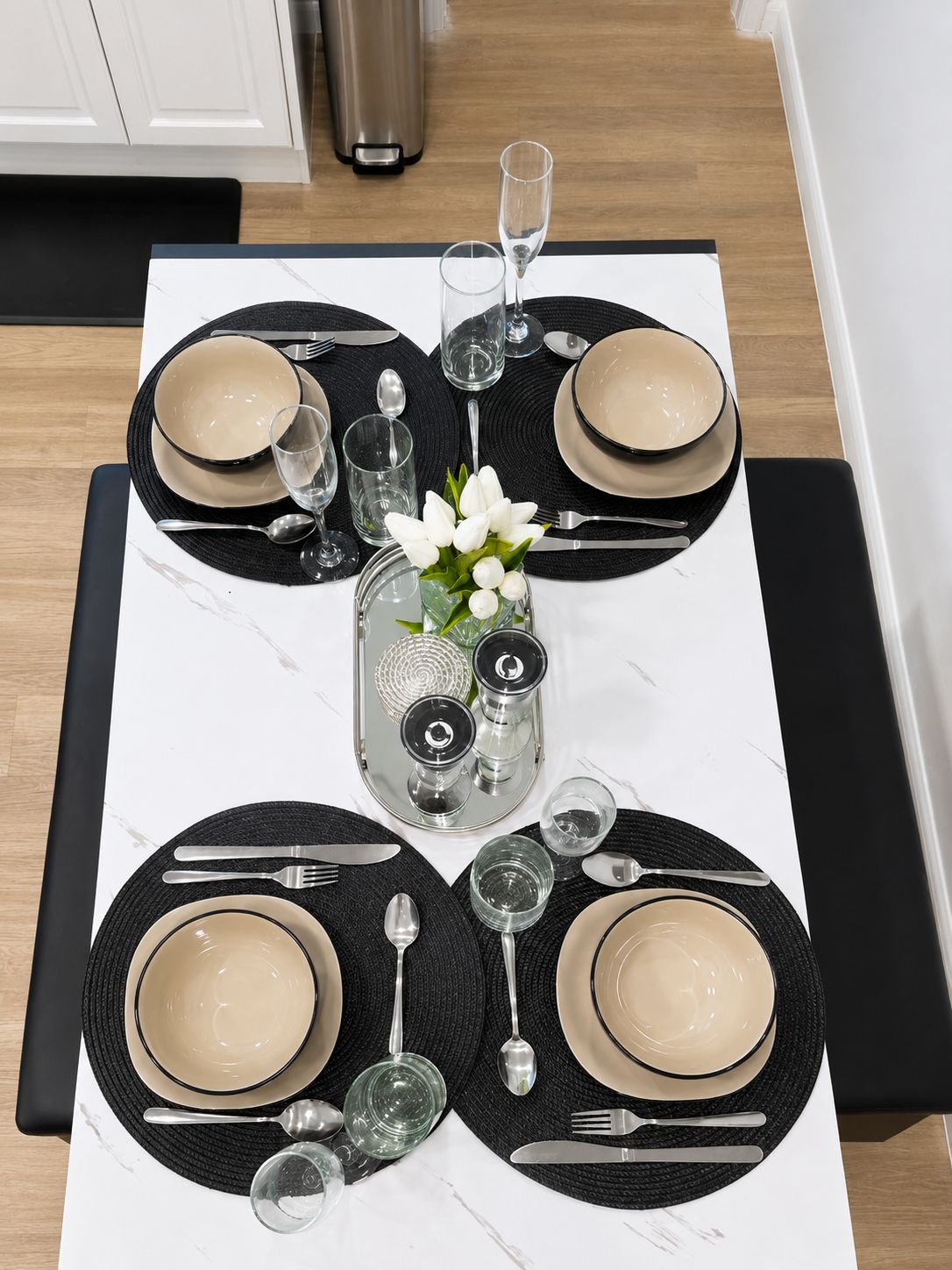 A dining table set with black placemats, beige plates, and silverware, centered with a glass vase of white flowers.