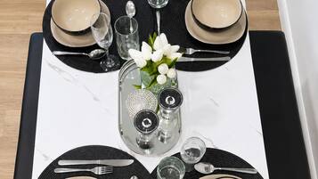A dining table set with black placemats, beige plates, and silverware, centered with a glass vase of white flowers.