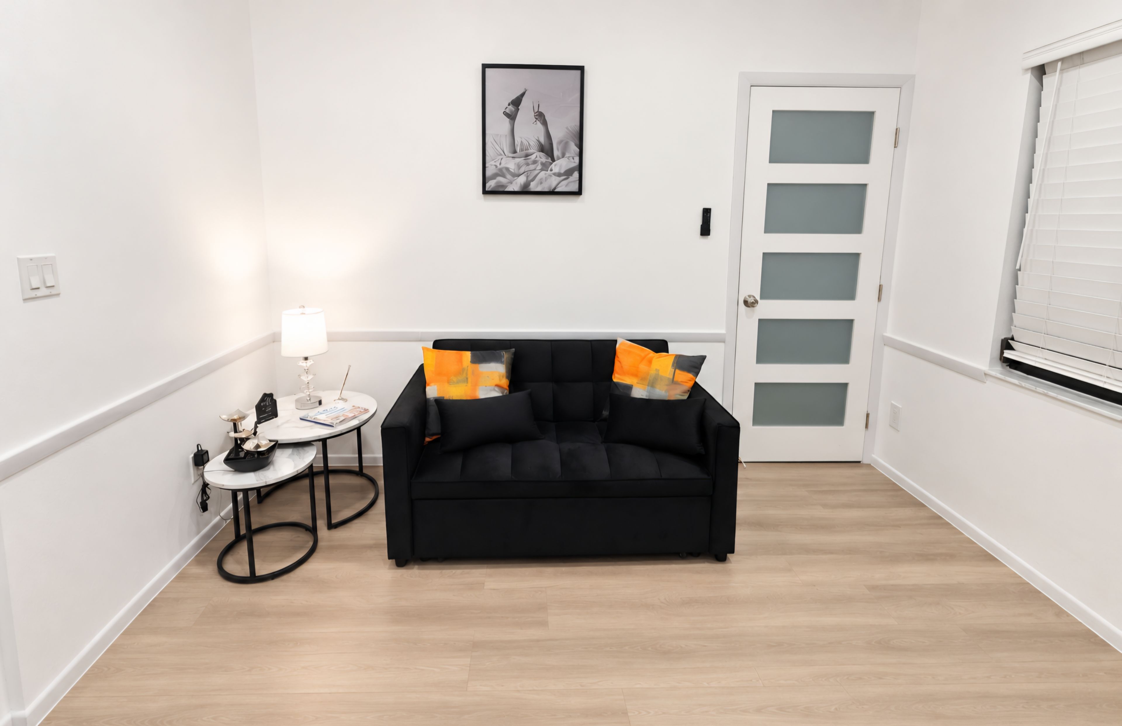 A modern living room with a black sofa, a small round table with a lamp, and a framed picture on the wall.