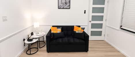 A modern living room with a black sofa, a small round table with a lamp, and a framed picture on the wall.