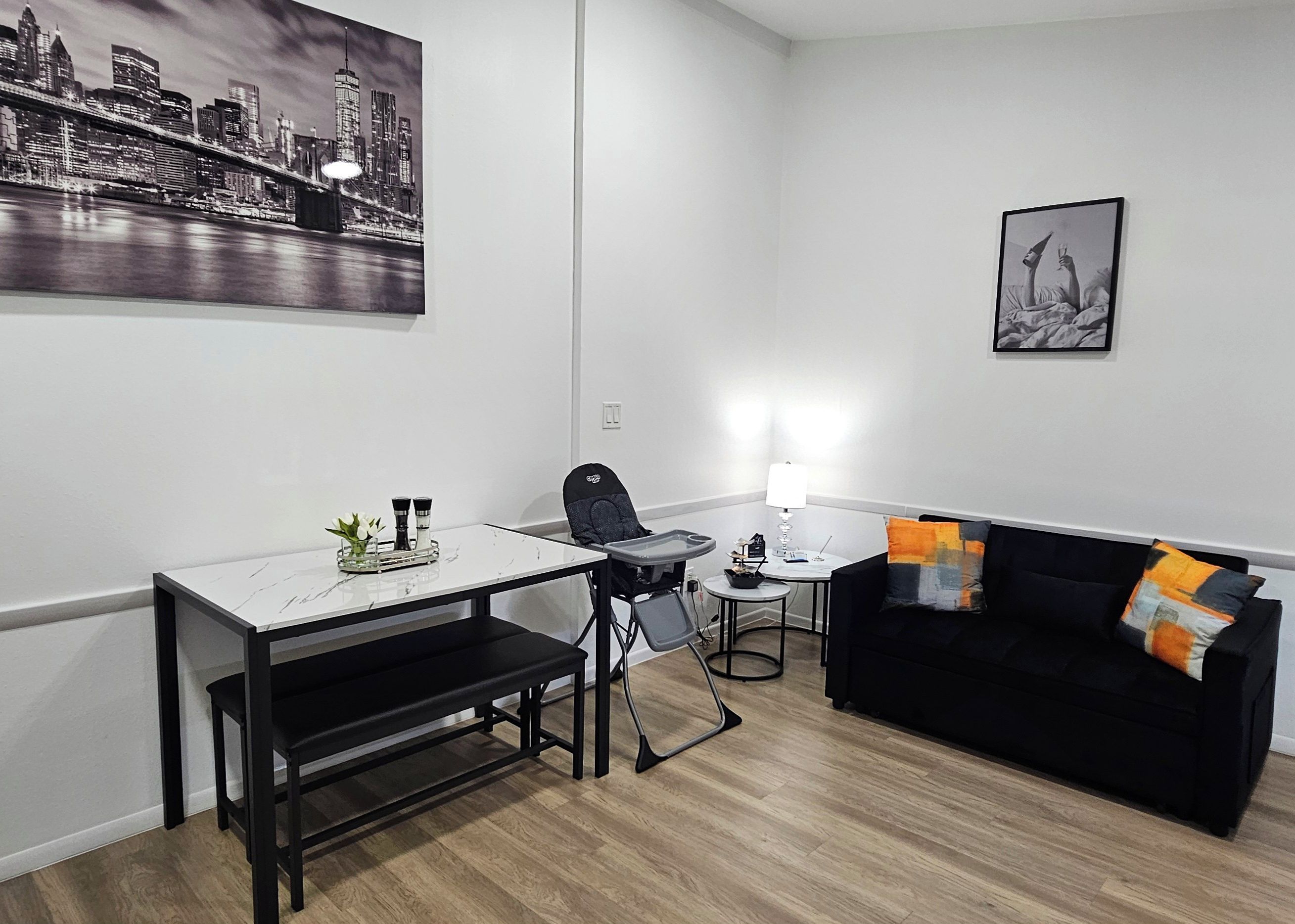 A modern living room with a black couch, a dining table with chairs, a small side table, and a cityscape painting on the wall.