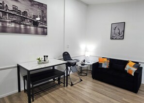 Un salon moderne avec un canapé noir, une table à manger avec des chaises, une petite table d’appoint et un tableau représentant un paysage urbain accroché au mur.